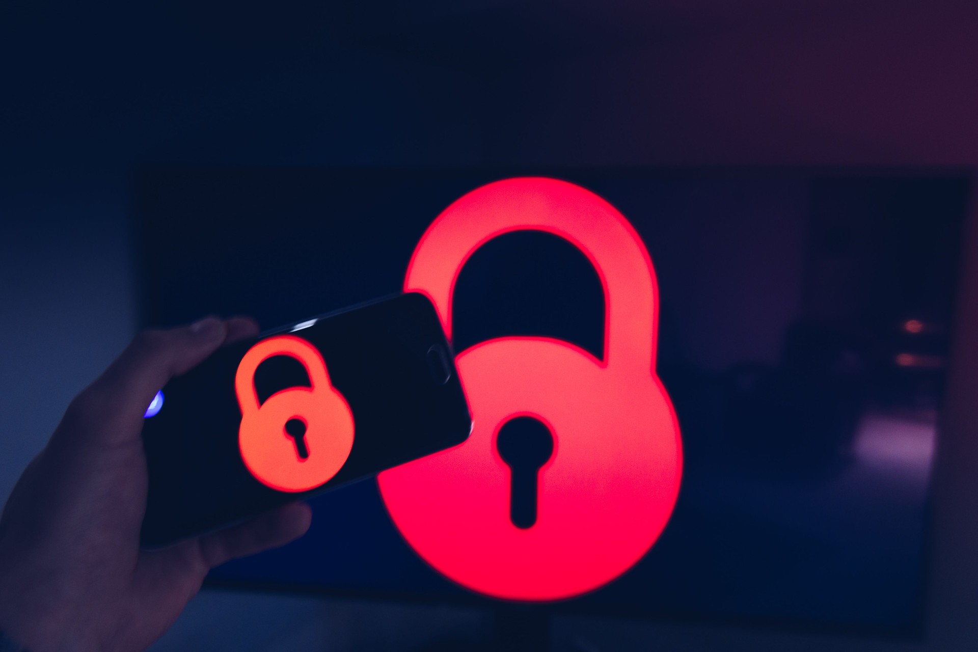 Cropped Hand Of Man Holding Mobile Phone With Lock Icon Against Monitor At Home